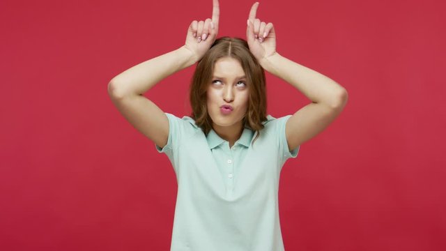 Positive playful young brunette woman in polo t-shirt standing with bull horn gesture and pretending to attack or defence, laughing, mooing like cow. indoor studio shot isolated on red background