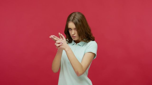 I'll kill you! Dangerous purposeful young woman in polo t-shirt holding gun gesture and searching for target, shooting at camera with finger pistol. indoor studio shot isolated on red background