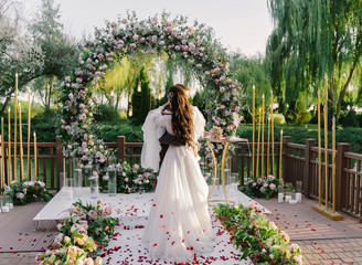 Newlywed bride and groom hugging and kissing at the wedding ceremony on the background of round wedding arch decorated with roses and eucalyptus leaves, guests are tossing red roses petals at them.