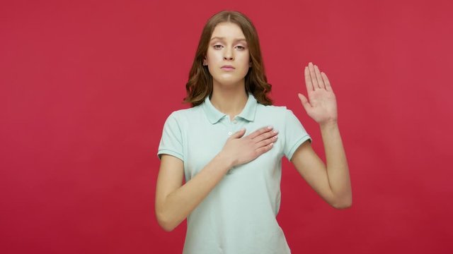 I swear to be honest! Sincere responsible young brunette woman in polo t-shirt raising hand to promise, taking vow with serious dedicated expression. indoor studio shot isolated on red background
