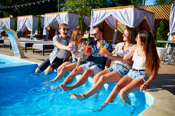 Obraz na płótnie Canvas Group of friends having fun at poolside party clinking glasses with fresh cocktails sitting by swimming pool on sunny summer day. People toast drinking beverages at luxury villa on tropical vacation.