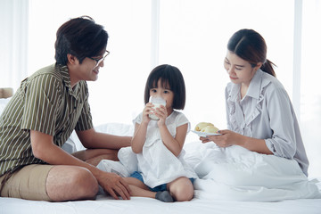 Happy Asian family having breakfast with milk bread sausages at bedroom, a little daughter drinking a glass of milk on bed at home