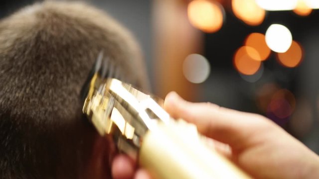 Trendy barber cuts bearded man's hair with a clipper in barbershop. Men's hairstyling and hair cutting in salon. Grooming the hair with trimmer. Hairdresser doing haircut in retro hair salon.