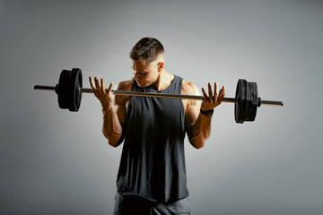 Fototapeta na wymiar A man does exercises with a barbell on a gray background. The athletic body of a young man in muscle tension makes an approach with a barbell. Copy space