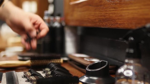 Barber takes modern wireless clipper from charging station in barbershop. Man's hand picks up trimmer from charger. Hair styling and grooming tools on the table in studio: brushes, scissors, combs.
