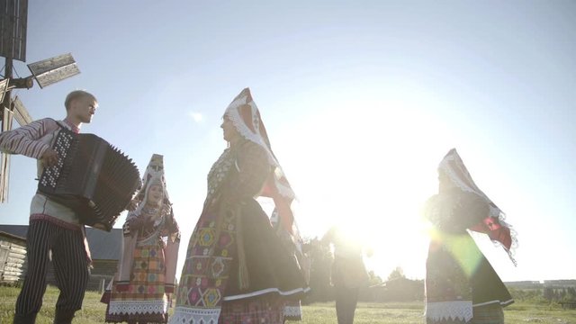 Folklore ensemble indigenous people dancing in traditional clothing outdoor on traditional antique wooden windmill background. Group of happy people wearing national Finno Ugric clothes at sunny day.