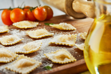 Tasty raw ravioli with flour, cherry tomatoes, sunflower oil and basil on a light wooden background. The process of making Italian ravioli.