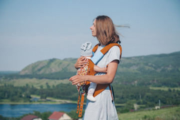 a woman with a child in a special carrier, a kangaroo in a walk. Village houses, the forest and river as the background. The concept of summer, warmth, freedom, village life, sunburn, childhood