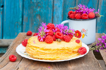Stack of freshly baked crepes with handful of fresh raspberries on top. Old utensils and field flowers on wooden table.