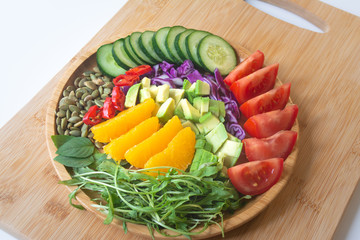 A buddha bowl of fresh summer healthy rainbow salad with cucumbers, tomatoes, arugula, basil, pumpkin seeds, avocado, red cabbage and chilli pepper on a wooden surface