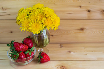 A bouquet of yellow dandelions on a warm wooden background. red strawberries in a glass vase. No one