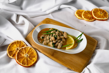 Marinated white beans in their own juice on an oval plate with Thai pineapple sauce and green onions. Decorated with a slice of lemon and dried oranges on a wooden Board with a snow white fabric