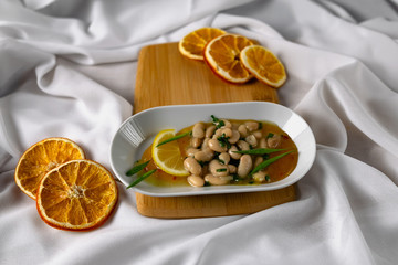 Marinated white beans in their own juice on an oval plate with Thai pineapple sauce and green onions. Decorated with a slice of lemon and dried oranges on a wooden Board with a snow white fabric