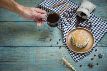 Hand held americano in clear cup with fresh bread put on wooden plate on black scottish pattern fabric,sack of roasted coffee beans,wooden spoon on old wooden table background.Top view with copy space