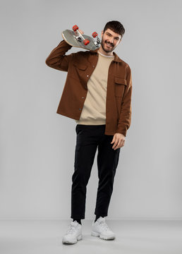 Leisure And People Concept - Happy Smiling Young Man In Brown Jacket And Black Pants With Skateboard Over Grey Background