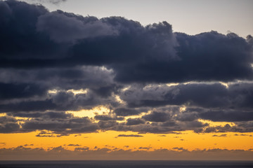 Aerial view of dark clouds back-lit by orange evening light over Tasman sea horizon