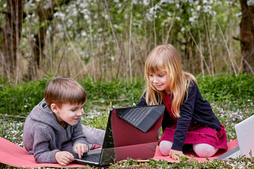 A girl and a smiling boy are sitting at laptops and studying in a flowered garden. Against the background of green grass and flowering trees. Remote entertainment.