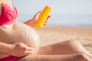 Sun shape created from sunscreen lotion on big belly. Young woman in pink swimsuit sitting on sand...