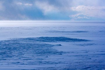 Blizzard on Lake Baikal in winter. Strong wind and a lot of snow. Blue shade of snow. Clouds in the sky. Horizontal.