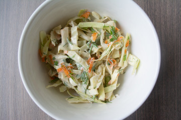 Healthy coleslaw salad with cabbage, carrots, dill and white dressing sauce in a white bowl on wooden surface