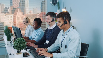 Multiracial call center agents talking to the clients in headset. There are skyscrapers in the...