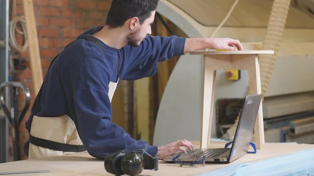 Young furniture maker skillfully make a wooden chair on a workbench and use laptop while working alone in his large woodworking shop. Handicraft and carpentry concept.