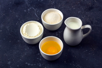 Baking background. Ingredients for cooking cake (flour, egg, sugar, milk) in bowls on dark table. Food concept. Close up