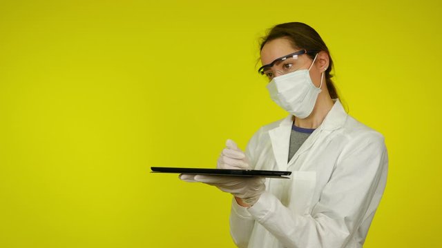 Woman in medical coat and protective mask uses holographic technology of the future on tablet screen. Augmented reality concept. Yellow background with copy space. Close-up. 4K footage