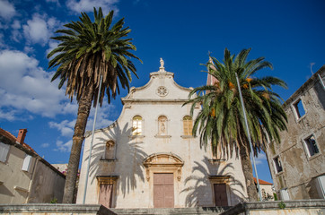 Fototapeta na wymiar Croatian seaside town, Dalmatia, medieval Roman Catholic church with palm trees with blue sky in summer sunshine.