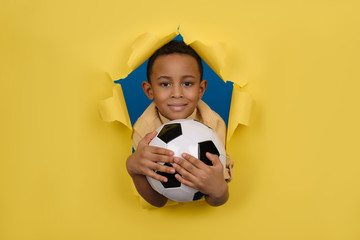 Smiling African-American boy soccer player and soccer fan in yellow Polo holds soccer ball in his hands against yellow torn paper wall background with space for text.