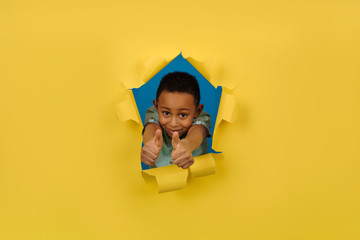 African-American boy on yellow background of torn paper shows positive hand sign, finger up or thumb up, gesture of approval or like, showing bursts of positive emotions. Concept of recommendation
