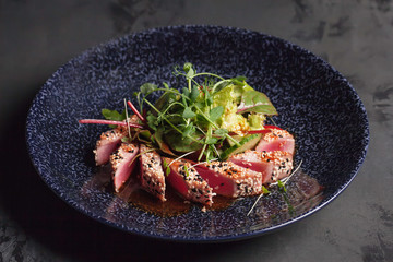 Restaurant menu. Salad with tuna, cucumber, herbs and micro greens. With sauce. Red tuna with sesame seeds. The blue plate. Red, green, blue. Dark background