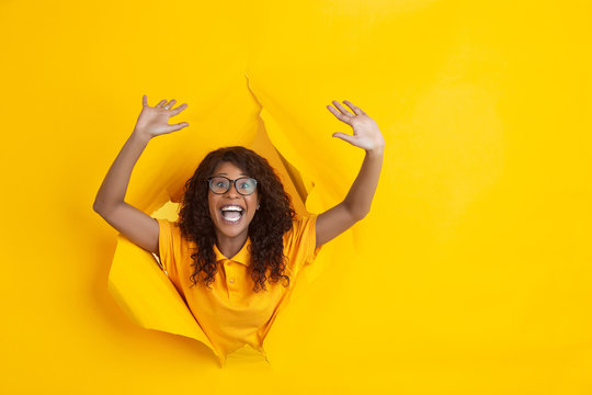 Crazy happy greeting. Cheerful african-american young woman in torn yellow paper background, emotional, expressive. Breaking on, breakthrought. Concept of human emotions, facial expression, sales, ad.