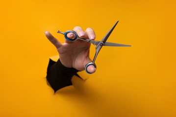 hairdresser holding in hand scissors for cutting hair from a torn hole in orange paper - obrazy, fototapety, plakaty