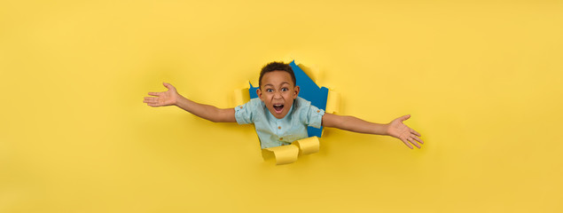 Cheerful and happy African-American on torn yellow paper wall background reaching out to support or take something asks to be picked up or hugged, boy asks to be picked up joyfully waving his arms.