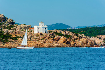 A view of a sailboat while sailing in the Mediterranean sea with the coast and a white lighthouse in the background on a sunny day with the blue sky on summer, in Sardinia Italy