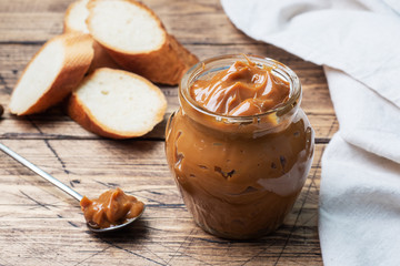 Boiled condensed milk with a glass jar on a wooden background. Sweet paste spread on slices of...