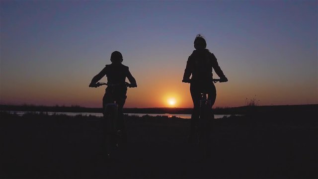 Silhouettes of two members of family - mother and son - riding their mountain bicycles near lake in amazing rays of sunset in slow motion. Sport and healthy lifestyle concept