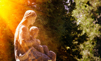 Fragment of ancient stone statue of Virgin Mary with the baby Jesus Christ in sonlight against green background of trees. - obrazy, fototapety, plakaty