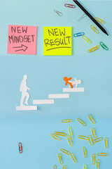 Top view of sticky notes with new mindset and new result lettering with paper clips, pencil and decorative men on career ladder on blue