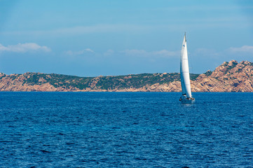 A view of a sailboat while sailing in the Mediterranean sea with the coast in the background on a day with the sun on summer, in Sardinia Italy
