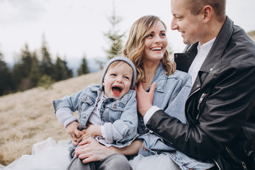 Stylish family in the autumn mountains. A guy in a leather jacket and a young girl in a gray-blue wedding dress with their son sitting on the grass together on the background of forest and landscapes