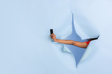 Female hand giving smartphone in torn blue paper hole background. Breaking on, breakthrought with gesturing. Concept of celebration, shopping, proposal, sales, ad. Copyspace. Promotions, discounts.