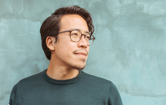 Portrait of an asian guy, wearing eyeglasses, standing in front of a light blue wall