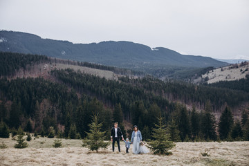 Stylish family in the autumn mountains. A guy in a leather jacket and a young girl in a gray-blue wedding dress with their son walking along the slope in the background of forest and landscapes