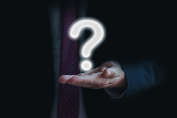 Businessman on dark blurred background holding question mark on his palm. Asking for answer or help