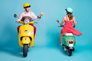 Full body photo of interested guy driving vintage moped blink opposite riding lady flirty handsome meet pretty girl direct finger wear retro clothes headgear isolated blue color background