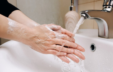 Two female hands with soapy foam in a stream of tap water in the bathroom. The concept of hygiene,...