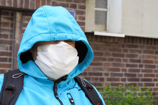 a light-skinned man in a blue jacket, hood, backpack and white face mask against the background of a brick house in the afternoon .during the coronavirus pandemic in Evropa .mask mode