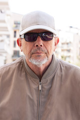 Casual dressed middle age or elderly man with black sunglasses, white cap, wearing windbreaker and a little gray beard looking straight into the camera. Close-up backlit image. Hillbilly portrait.